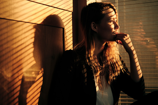 Portrait Of Pensive Business Woman Standing By Window With Venetian Blinds Projecting Shadows At Sunset