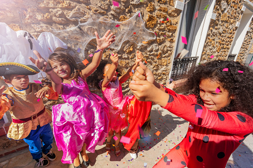 Group of diverse looking kids throw confetti wearing Halloween carnival costumes having fun jumping and lifting hands