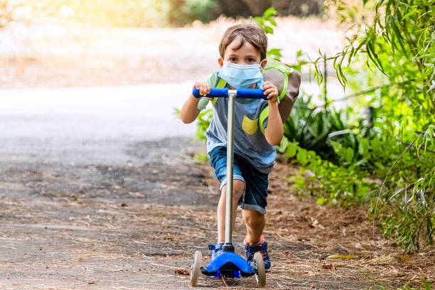 istock Little boy riding a push scooter carrying a back pack wearing a protective face mask 1253176395