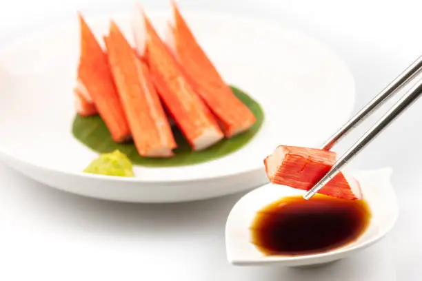 Photo of Stainless steel chopsticks are squeezing Crab Stick Sashimi in a white dish with dipping sauce with tamari shoyu and wasabi.