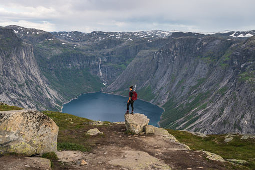 Asian woman traveller with backpack standing on rock and enjoying landscape of mountains and lake in Trolltunga mountain cliff trail, Odda town, Norway, Scandinavia, Europe