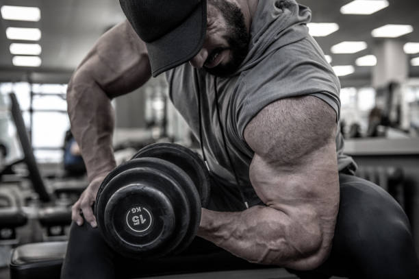 strong young bearded man in cap lifting heavy weight dumbbell on muscle biceps sitting with great effort and pain during hard workout training in sport gym - doping imagens e fotografias de stock