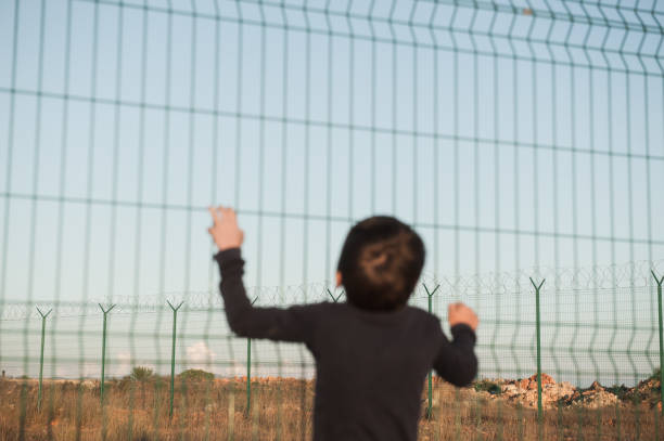 juvenile delinquent little refugee ilegal immigrant kid in displaced persons camp with high fence with barbed razor wire with hope of freedom in Mexico USA Europe Turkey Syria juvenile delinquent little refugee kid in displaced persons camp with high fence with barbed razor wire with hope of freedom deportation stock pictures, royalty-free photos & images