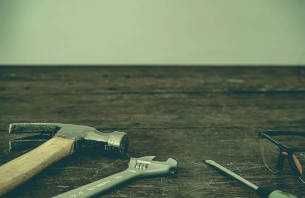 Hand tools include a hammer, sliding wrench, Screwdriver, and safety goggles are placed on a vintage old brown wooden table.