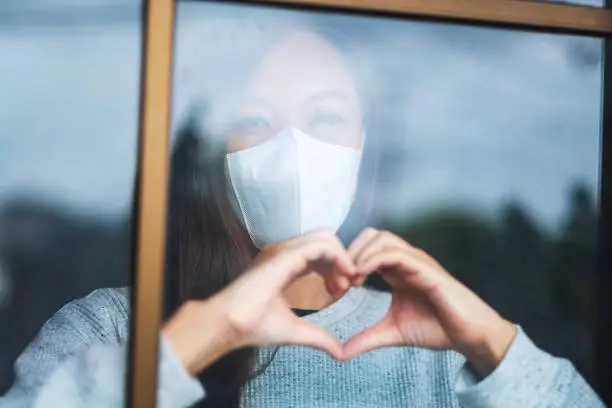 A young asian woman wearing protective mask doing heart hand sign while staying isolation at home for self quarantine and COVID-19 concept