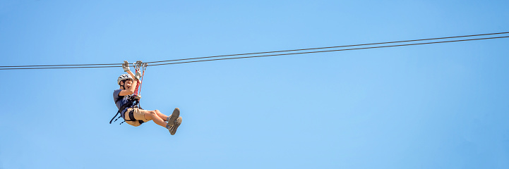 Teenager having fun on a zipline on panoramic blue sky background with copy space.