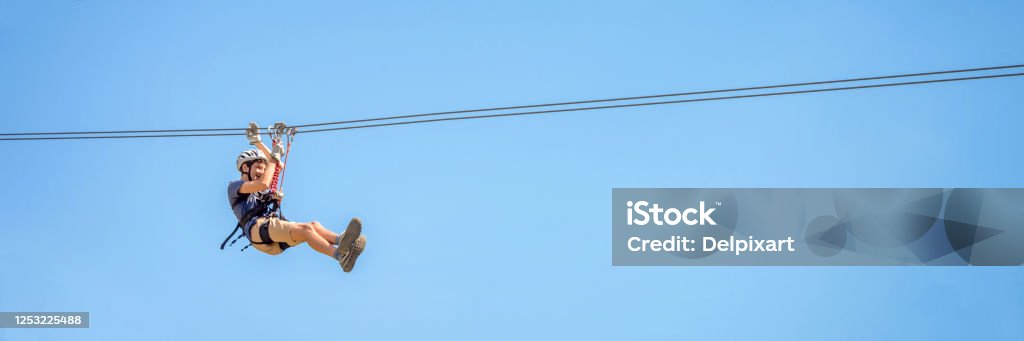 Teenager having fun on a zipline on panoramic blue sky background with copy space. Zip Line Stock Photo