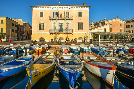 Bardolino, Verona / Italy - Feb 28 2020: Colored fishing boats docked in the little harbor with the town hall in the background