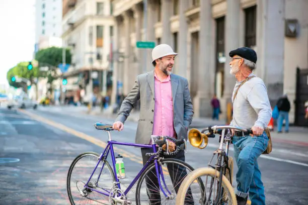 Mature and senior male friends in casual clothing standing with their bicycles and talking in middle of open street in downtown Los Angeles.