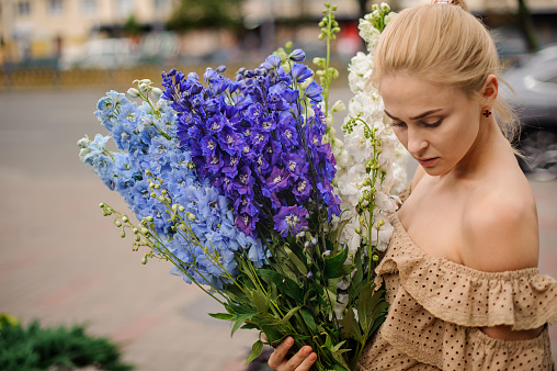 young blonde woman holds large bouquet of white and blue colors delphinium in her hands and looks down