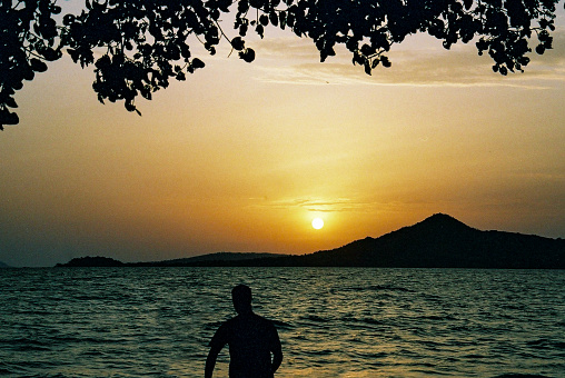 Lake Victoria Sunset towards Rusinga Island Kenya.  Silhouette of a man fishing looking at the sunset with a tree branch silhouette as framing above.