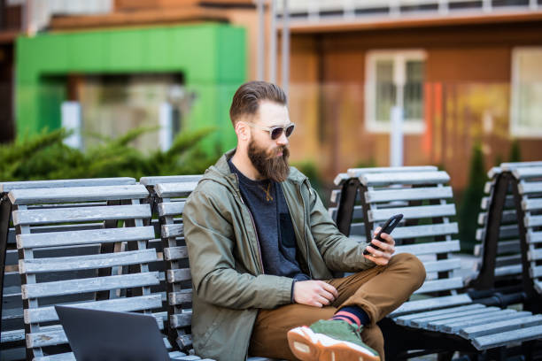 istock Young bearded hipster man sitting on the bench and use phone outdoors 1253242529