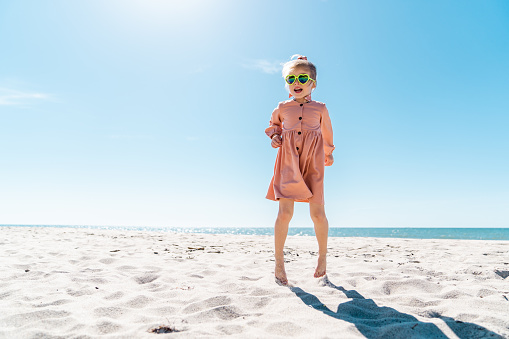 Beautiful little girl wearing beige dress is running and jumping on the empty beach. Sea and cloudless sky on the background.