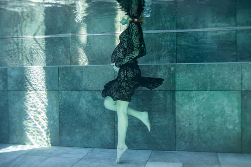 Pregnant woman in black dress swimming and diving in swimming pool and looking in the mirror. Underwater view