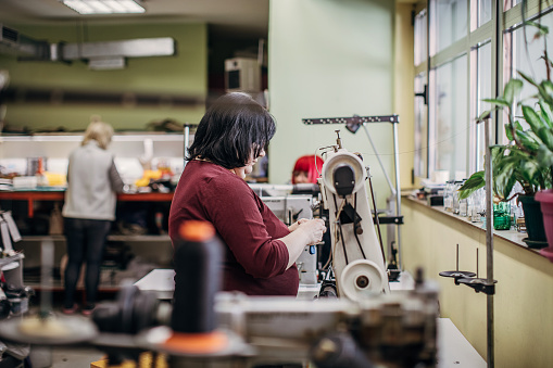 One mature woman working on sewing machine in workshop, making female leather purses.