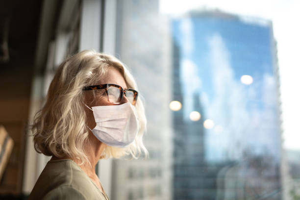 Mature businesswoman looking out of window with face mask Mature businesswoman looking out of window with face mask new normal concept stock pictures, royalty-free photos & images