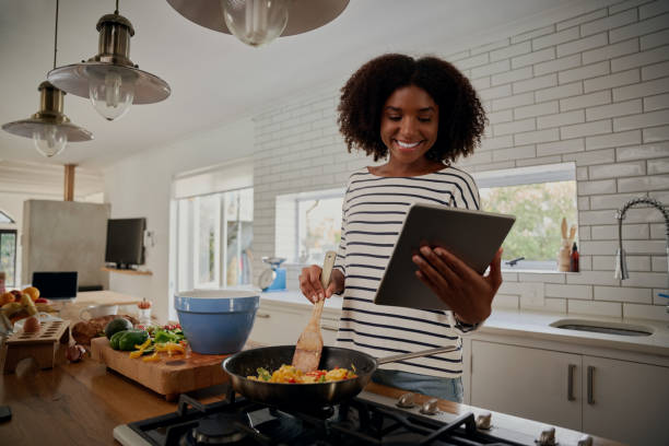 joven mujer africana viendo la receta en la tableta digital mientras cocina el almuerzo en la cocina moderna - cocinar fotografías e imágenes de stock
