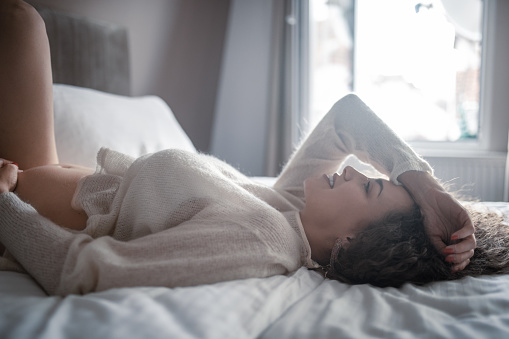 Beautiful young pregnant woman is touching pregnant belly while lying on the bed in bedroom and enjoying morning lights