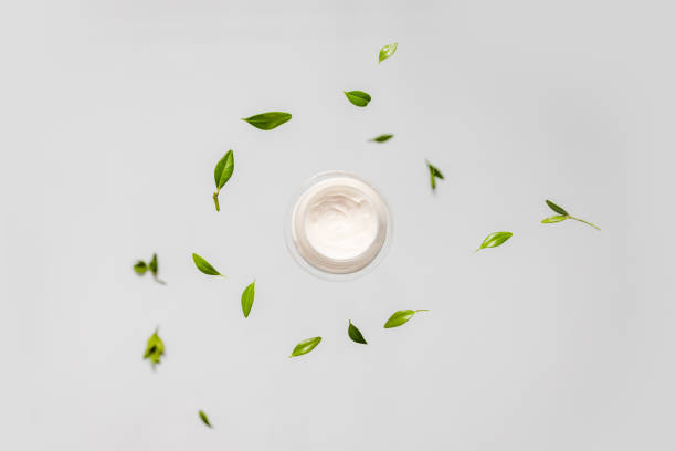 istock Fresh green leaves falling and cream jar on a grey background. Flat lay, top view, copy space 1253250545