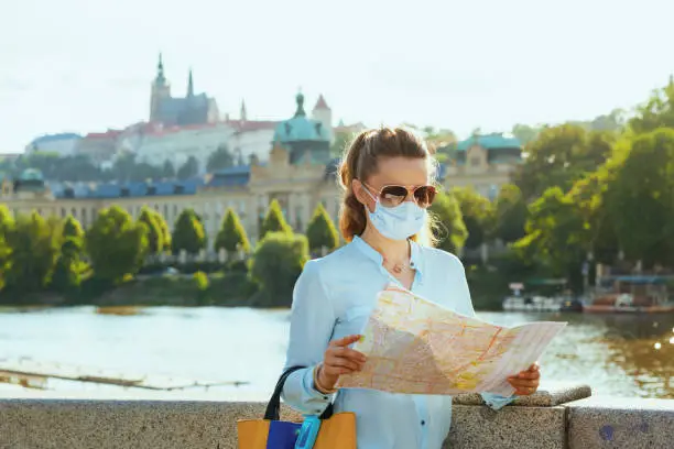 Life during coronavirus pandemic. elegant solo tourist woman in blue blouse with medical mask, sunglasses and map sightseeing outdoors on the city street.