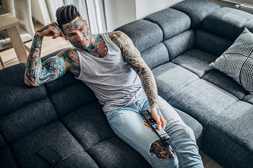 One young modern man with whole body covered in tattoos sitting on sofa in his living room and watching TV.