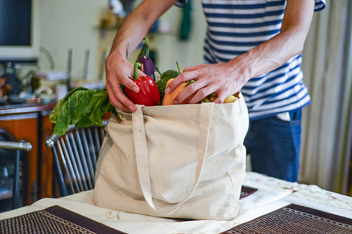 Asian man unpacking groceries at kitchen island. He is removing fruits and vegetables from reusable bags.