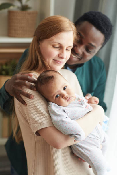 Happy Mixed-Race Family at Home Vertical portrait of young interracial family holding cute mixed-race baby while embracing lovingly standing by window at home biracial newborn stock pictures, royalty-free photos & images