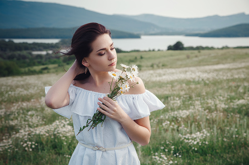 Beautiful young brunette woman in a white dress is enjoying spring in a field of daisies. Health idea and concept, beautiful healthy hair and skin, lack of allergies