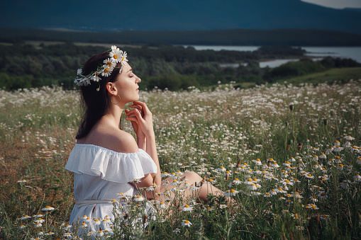Beautiful young brunette woman in a white dress is enjoying spring in a field of daisies. Summer countryside concept. Close to nature vacation. Woman and daisies. Summer light