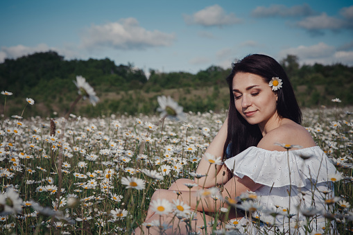 Beautiful young brunette woman in a white dress is enjoying spring in a field of daisies. Health idea and concept, beautiful healthy hair and skin, lack of allergies