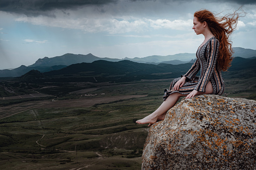 Ginger woman sits on a cliff and looks at an impending storm, watching the last light. Hair blowing in the wind.Girl admiring mountain landscape. Crimea, Mount Klimentieva