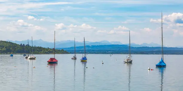 Ammersee, Bavaria / Germany - June 3, 2020: Panorama with colorful sailboats anchoring at Ammersee (Lake Ammersee). Alps on the horizon.