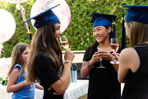 Celebrating primary school graduation in the backyard during Covid-19 pandemic for three multi-ethnic teenage girls.They are drinking with graduation hats and non-alcoholic beverage. Horizontal outdoors waist up shot with copy space.