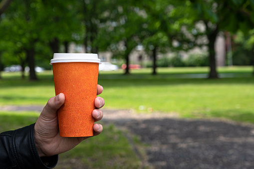 Close up of man hand holding a paper orange cup of coffee on park background