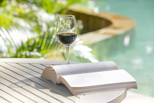 Open book and a glass of red wine on the table next to the swimming pool. Summer day leisure concept.