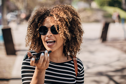 One beautiful happy woman with curly hair recording a voice message with smart phone on the street.