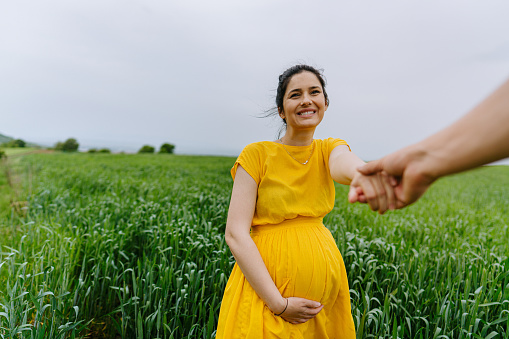 Photo of a smiling pregnant woman holding hands with her partner in the middle of a grass field, enjoying a beautiful spring day while expecting a new life to begin.