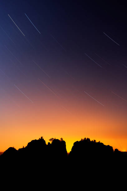 istock Silhouetted rocks and circumpolar stars. Beautiful night view over sandstone cliff into deep misty valley in Saxony Switzerland, colour background with orange and blue sky star sun on sky, Germany 1253293037