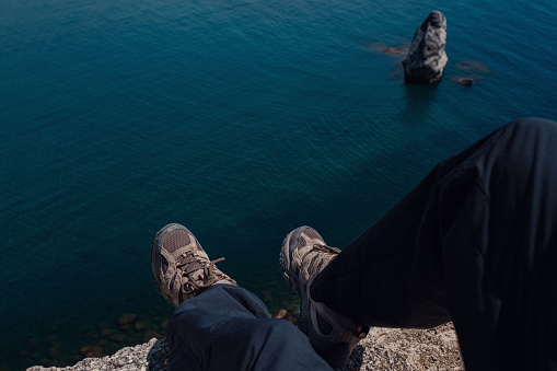 Close-up of hiking boots of an independent male traveler on top of a mountain, looking at the view on ocean in sunny day