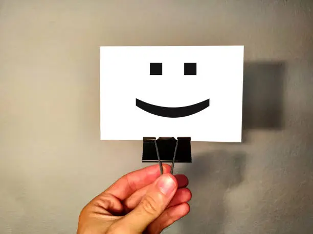 Photo of Male hand holds a white card on a background of a gray wall on a metal clothespin with a smile icon. Service rating, satisfaction concept.