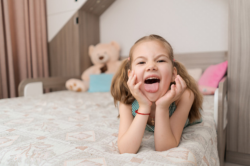 Lovely carefree kid portrait making sweet, funny faces while lying on her bed in her room