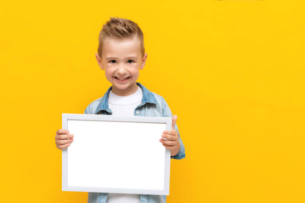 Happy smiling kid holding white frame with copy space for text certificate or diploma Happy smiling kid holding white frame with copy space for text certificate or diploma. Blank empty white frame in hands isolated on yellow background kids using smart board stock pictures, royalty-free photos & images