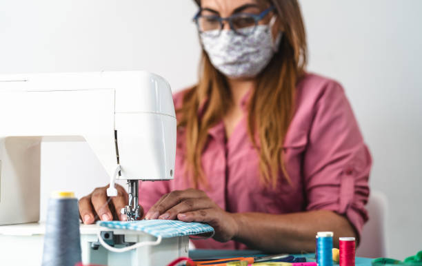 mujer madura que trabaja con la máquina de coser haciendo mascarilla facial médica casera para prevenir y detener la propagación del virus corona - concepto de costurera textil y personas de salud - seam needle textile industry thread fotografías e imágenes de stock