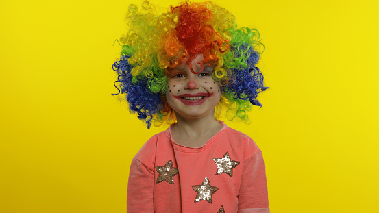 Child girl clown in colorful wig making silly faces. Happy five years old little caucasian kid having fun, smiling, dancing, looking at camera. Expressions. Halloween. Yellow background. Chroma Key