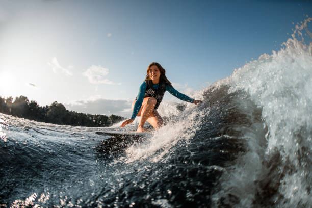 smiling young woman wakesurfing down the river waves smiling young woman in swimming vest wakesurfing down the river waves. Beautiful blue sky on the background river swimming women water stock pictures, royalty-free photos & images