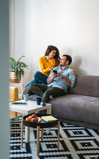 Man holding in hands cell phone and woman showing screen and smiling together while sitting on sofa at home