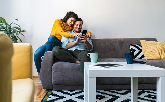 Man holding in hands phone and woman showing screen and smiling together embraced while sitting on sofa at home