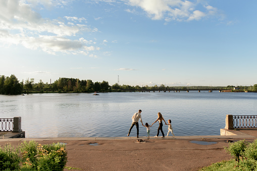 A young family walks hand in hand along the river. Daughters hold their parents ' hands and look at the water and blue sky with clouds. A family with children spends time in nature with clean air and blooming trees on a sunny summer day.