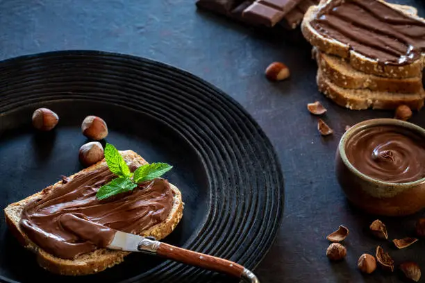 Chocolate cream spread on bread slice and hazelnuts on a dark black rustic plate with mit leaves on top and chocolate bars