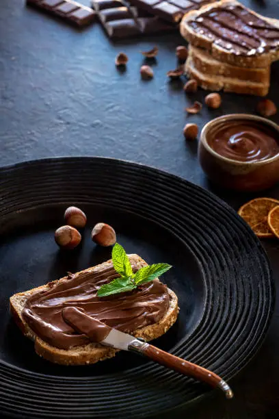 Chocolate cream spread on bread slice and hazelnuts on a dark black rustic plate with mit leaves on top and chocolate bars